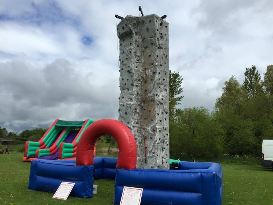 Grey climbing tower set up on grass beside a green and red inflatable slide, rules signs at the base