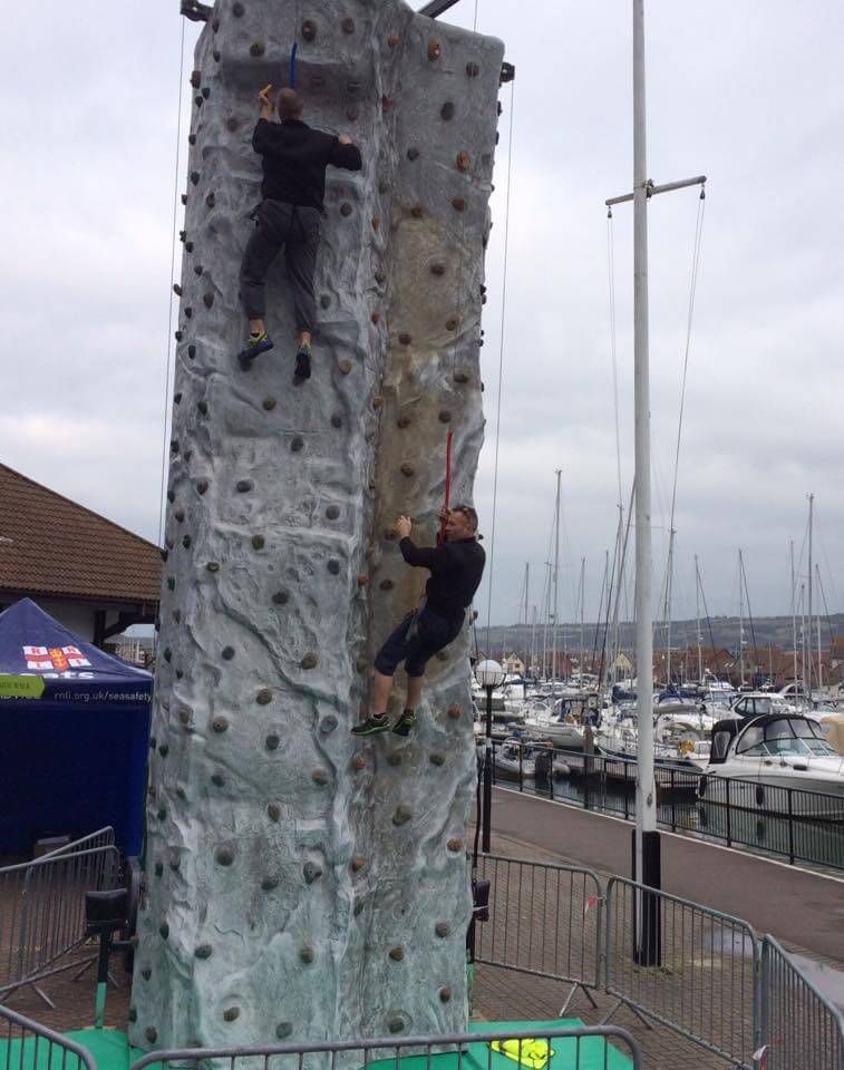 Two men climbing the grey wall behind metal barriers at a marina, yachts moored behind