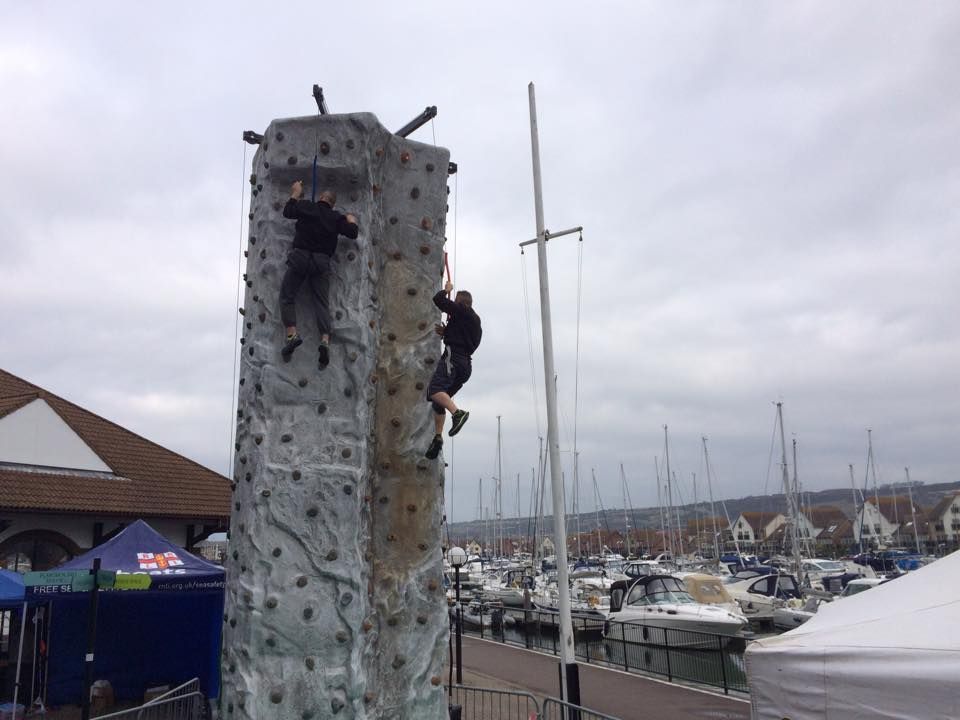Two people climbing the grey wall at a quayside marina, moored yachts and boats behind