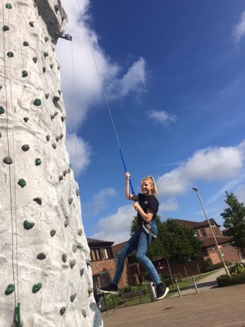 Smiling girl being lowered on the blue belay line beside the wall, brick building and trees behind