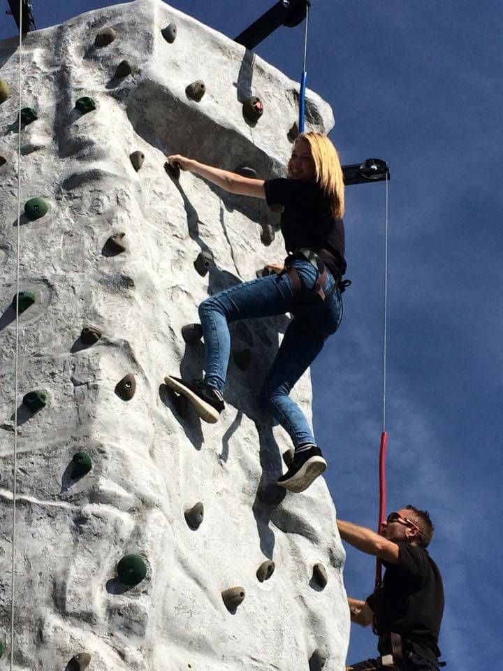 Blonde woman in a harness climbing the grey rock-effect wall against a blue sky, staff member below