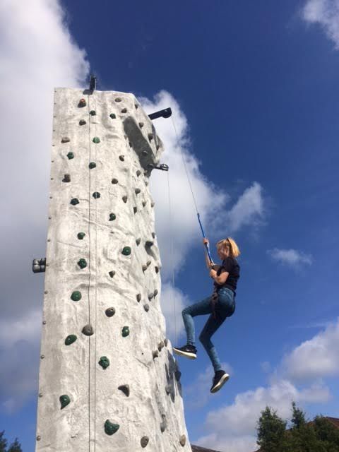 Climber near the top of the grey wall gripping the blue auto-belay line against a bright blue sky