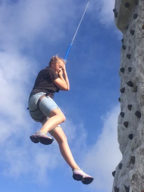 Girl in shorts dangling from the blue auto-belay line beside the climbing wall against a blue sky