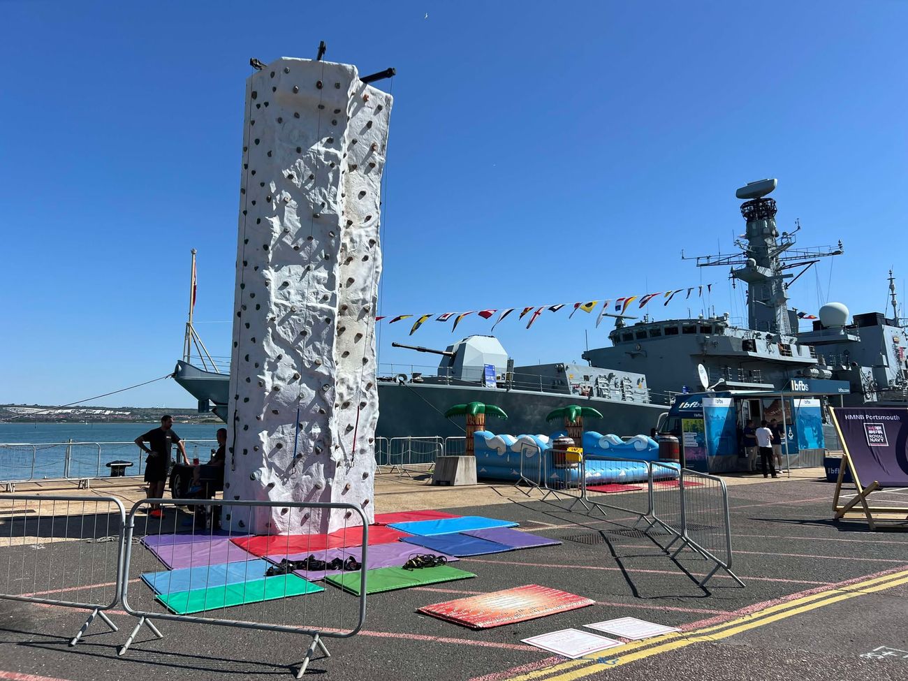 White climbing tower on the quayside at HMNB Portsmouth with a Royal Navy warship behind, crash mats at the base